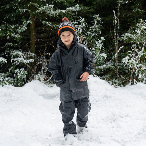 Boy in snow wearing a black waterproof jacket, pants, and orange pom pom hat.