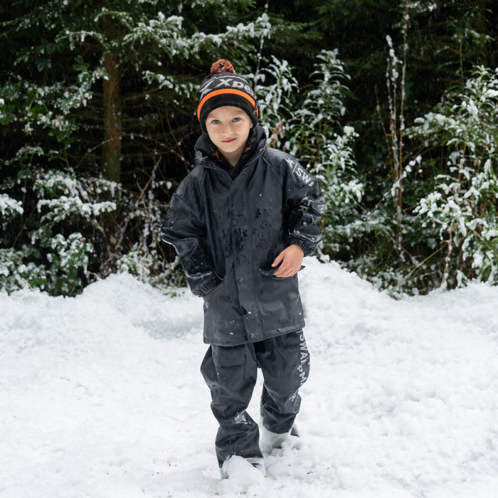 Boy in snow wearing a black waterproof jacket, pants, and orange pom pom hat.