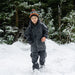 Boy in snow wearing a black waterproof jacket, pants, and orange pom pom hat.