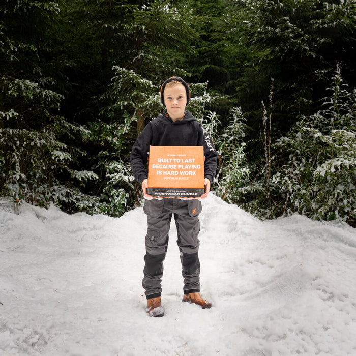 A boy in snow holds an orange box labeled "Built to Last Because Playing is Hard Work Workwear Bundle".